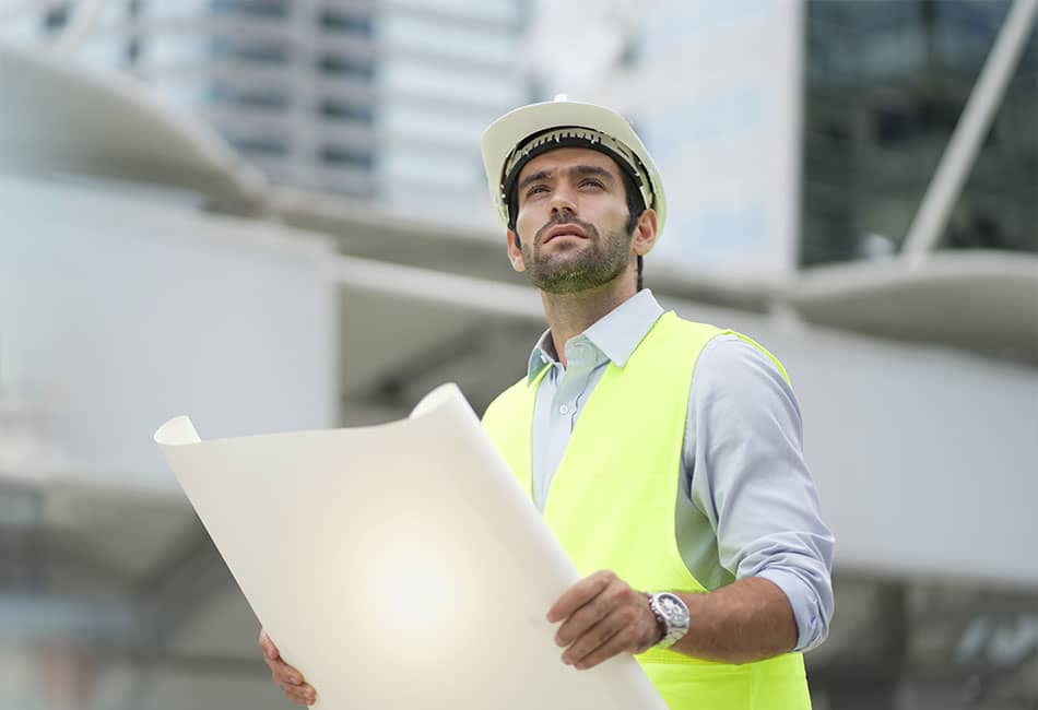 A man in a high-visibility vest holding a plan while looking at buildings.