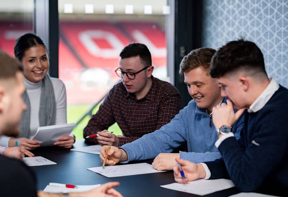 A group in a meeting around a desk with sports stadium stands visible in the background through a window.